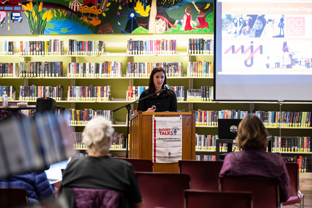 Lori Kido洛佩兹站在一个讲台在图书馆设置。她站在架子上的书前交付一个跟一个坐着的观众。
