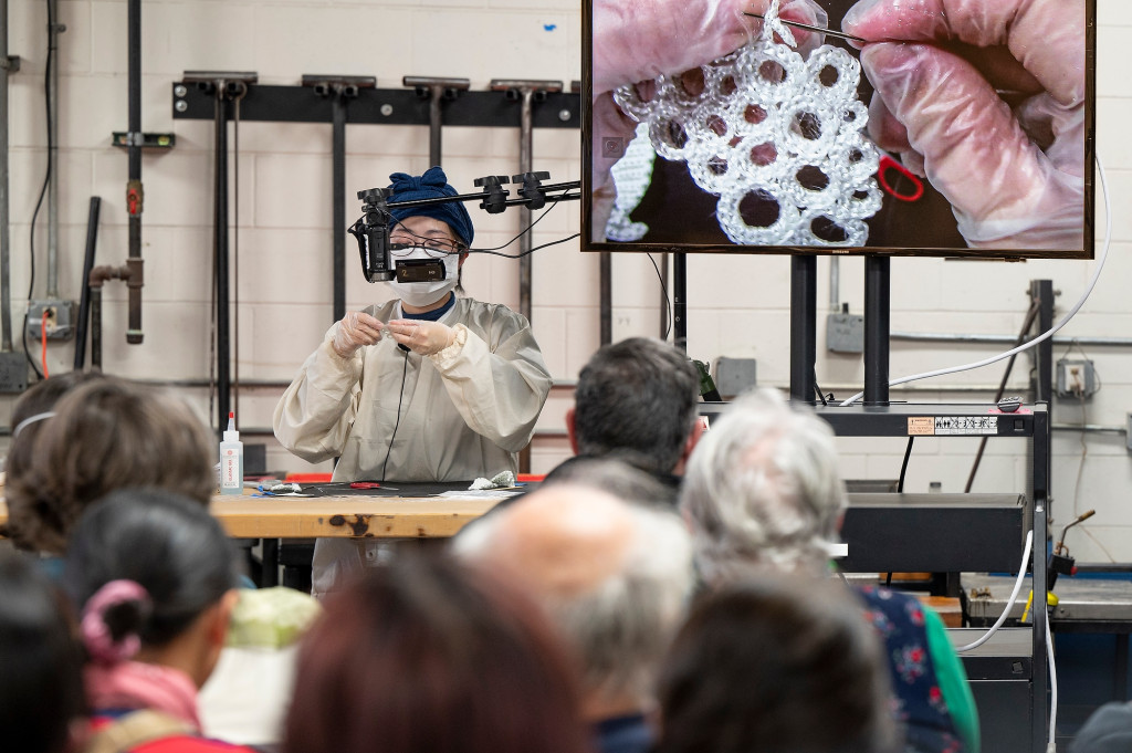Machiko Ito站在前面的玻璃实验室一个坐着的观众。她工作在一个钩针编织玻璃纤维圆,扩大对旁边的观众在屏幕上她的工作表。