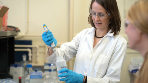 在实验室环境中,克里斯蒂Remucal穿着一件白色外套,防护眼镜和手套,用吸管了样品瓶。
