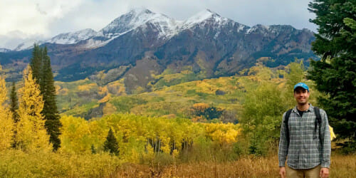 秋天的一天，福特·弗莱伯格站在落基山脉前，在照片的右下角显得很小。前景中的森林正由绿色变成金色。