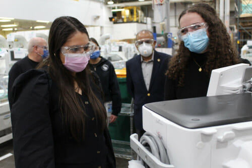 两名戴着医用口罩、穿着商务休闲装的妇女看着一台台式实验室设备旁边的显示器。背景中站着三个穿着相似的人。他们处于工业环境中。房间里似乎装满了设备。