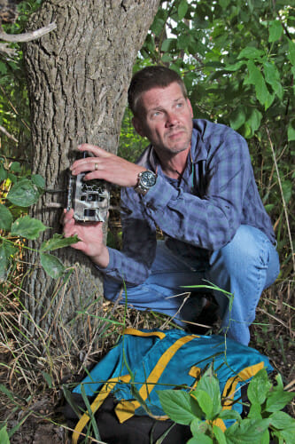 蒂姆·范Deelen蹲在树林里一个树干。他是定位跟踪相机在树上和展望未来沿着小路相机的视线。