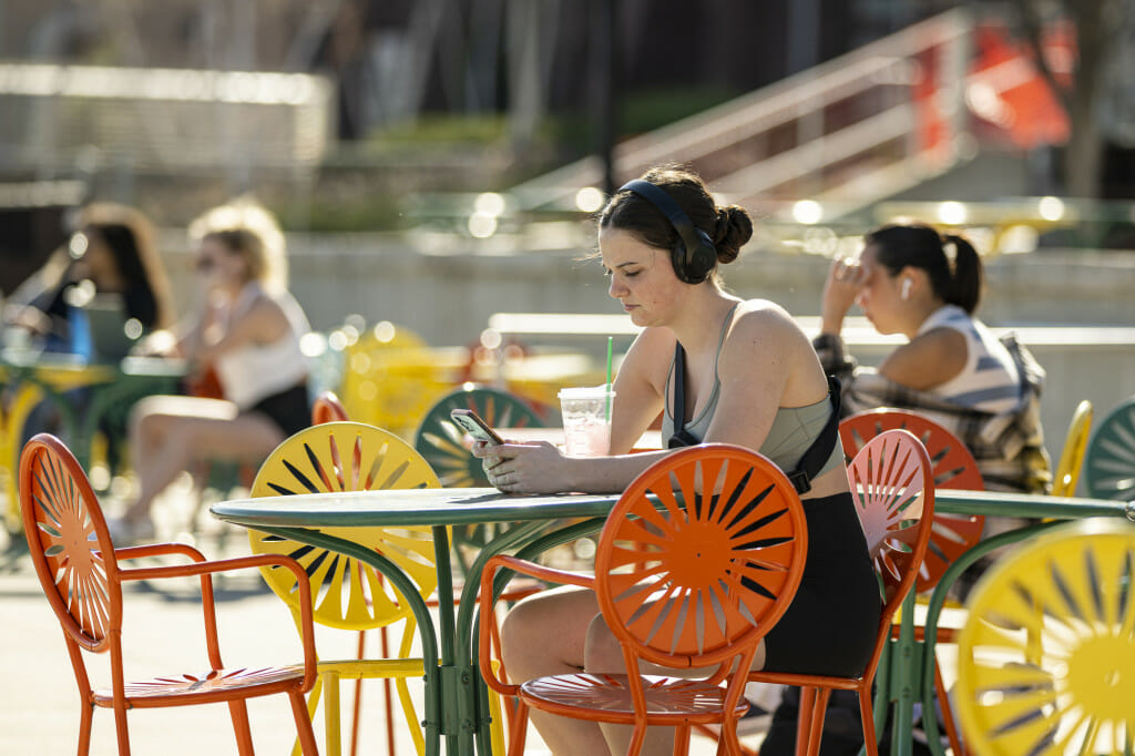 一个学生,萨米Lacke,戴着耳机,看着她电话坐在餐桌旁纪念联盟平台在一个阳光明媚的一天。