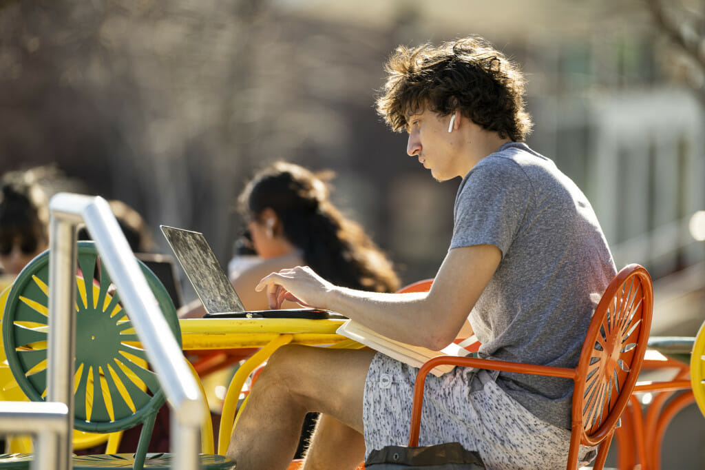 学生昆西油田工作穿空气舱并且保持打开一本书,坐在他的膝盖上,他的工作在他的笔记本电脑,坐在一张桌子上纪念联盟平台在一个阳光明媚的一天。