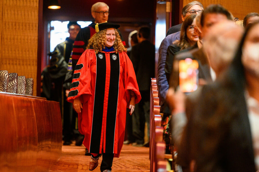 詹妮弗Mnookin,学术长袍,穿红色黑色博士的帽子,走进礼堂。托尼•埃弗斯州长还穿着学术徽章,走在她的身后。