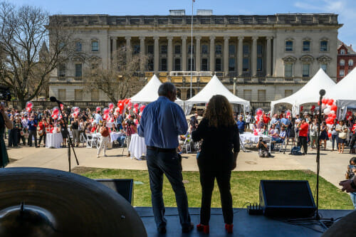 两个人站在一个阳光广场前的舞台上向那里人民讲话
